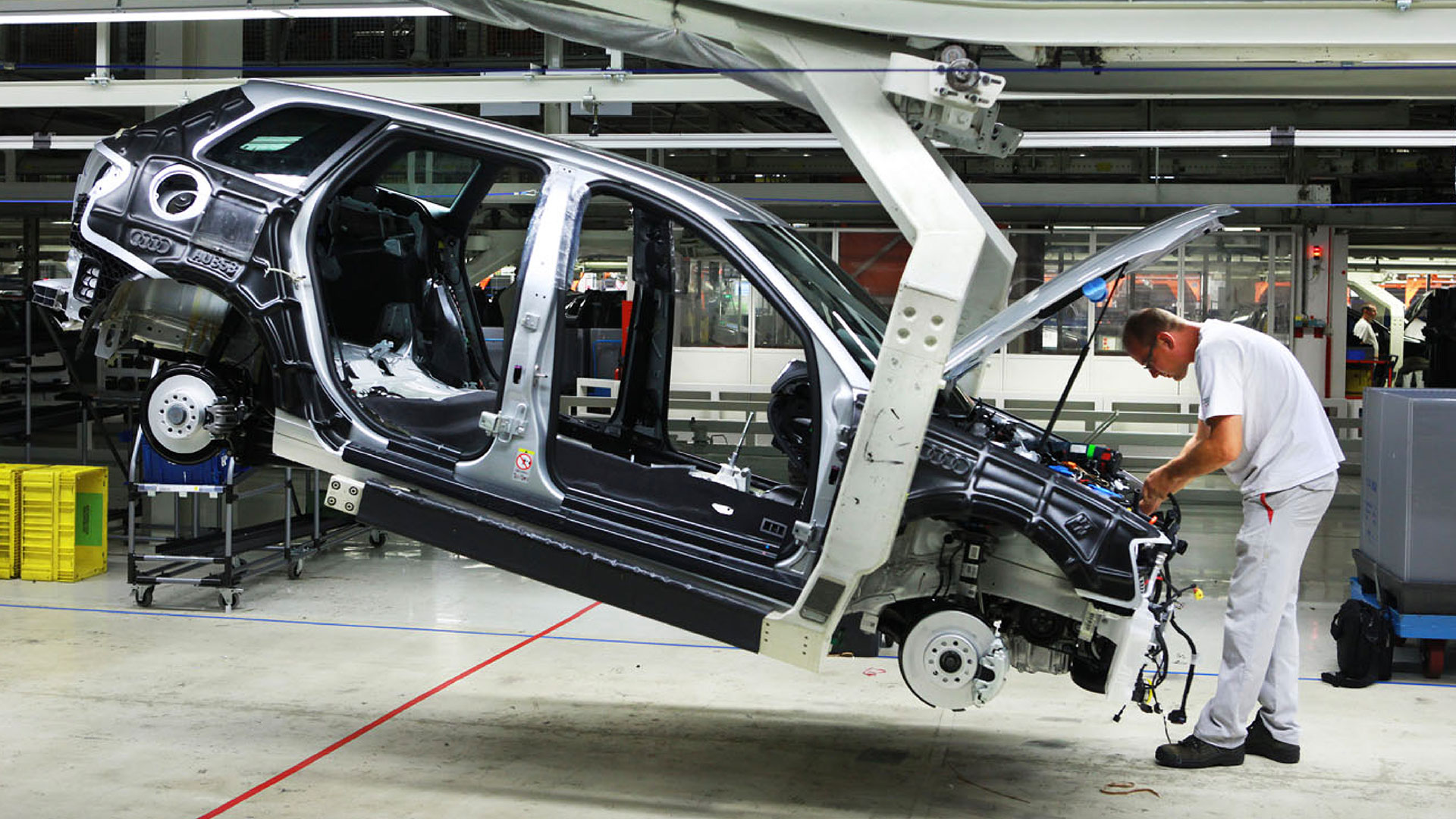 A worker assembling components under the car's hood with the help of a robot that holds the car and tilts it forward, making the worker's job easier